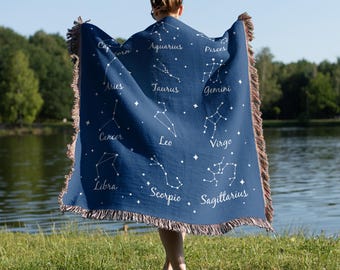 Könnte beinhalten: Eine marineblaue Decke mit weißen Sternbildern und Sternmustern, mit den Namen der Tierkreiszeichen. Die Decke hat einen Fransenrand in Braun- und Beigetönen. Eine Person hält die Decke in einer grasbewachsenen Gegend.