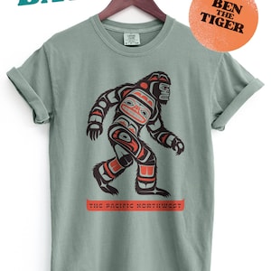 Puede incluir: Una camiseta verde salvia con un gr&aacute;fico estilizado de Sasquatch en negro, rojo y naranja. El texto "The Pacific Northwest" est&aacute; impreso debajo del gr&aacute;fico. El texto "Ben The Tiger" est&aacute; impreso en un c&iacute;rculo de color melocot&oacute;n en la esquina superior derecha.