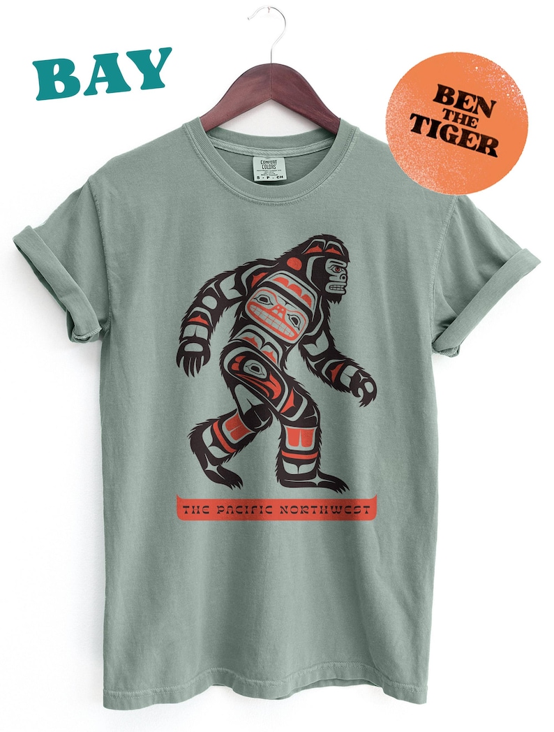 Puede incluir: Una camiseta verde salvia con un gr&aacute;fico estilizado de Sasquatch en negro, rojo y naranja. El texto "The Pacific Northwest" est&aacute; impreso debajo del gr&aacute;fico. El texto "Ben The Tiger" est&aacute; impreso en un c&iacute;rculo de color melocot&oacute;n en la esquina superior derecha.