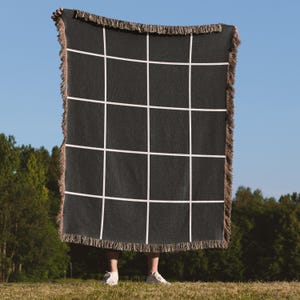 May include: A rectangular woven throw blanket featuring a dark charcoal gray base with a prominent white grid pattern. The edges of the blanket are finished with a textured, multi-colored fringe detail along all four sides of the fabric piece.