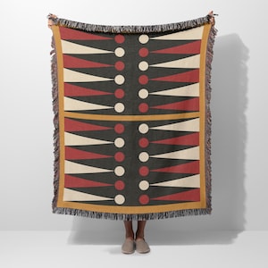 May include: A woven throw blanket with a geometric pattern resembling a backgammon board. The design features alternating red, cream, and black triangles and white circles. The blanket has a mustard yellow border and fringe.