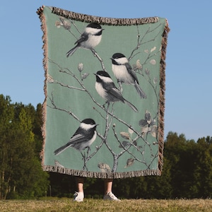 May include: A green throw blanket featuring a detailed illustration of several black-capped chickadees perched on tree branches. The blanket has a fringed edge and is held up against a blue sky background.