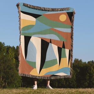 May include: A woven throw blanket featuring a geometric landscape design with abstract shapes in shades of terracotta, sage green, sky blue, mustard yellow, cream, and black. The rectangular blanket has a fringed edge on all four sides.