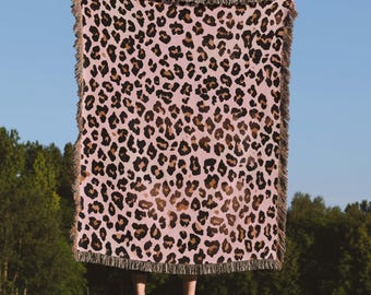 Op de afbeelding: Een roze sprei met een luipaardprint. De sprei heeft bruine en zwarte vlekken en een franjerand. De sprei wordt omhooggehouden tegen een blauwe lucht en groene bomen.