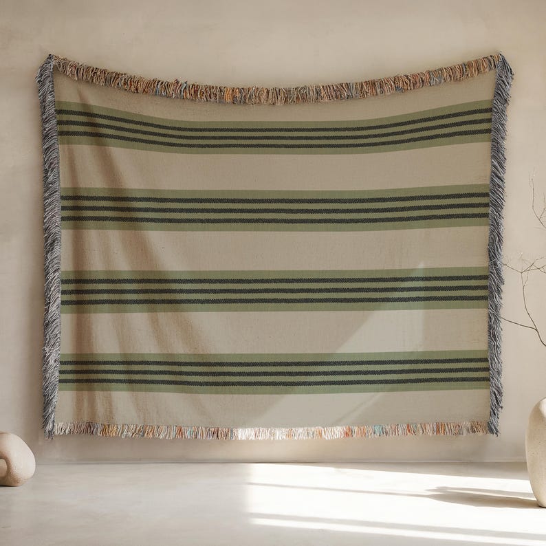 K&ouml;nnte beinhalten: Eine beige-gr&uuml;n gestreifte Decke mit Fransen. Die Decke hat horizontale Streifen in Gr&uuml;n- und Dunkelgraut&ouml;nen. Die Kanten sind mit einem mehrfarbigen Fransen versehen. Die Decke h&auml;ngt an einer Wand.