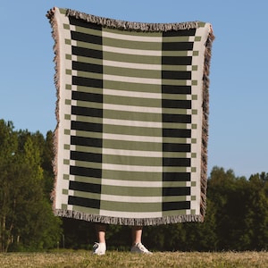 May include: A rectangular woven blanket with a geometric pattern of alternating black, olive green, and off-white stripes. The blanket has a fringed edge and is held up against a blue sky background.