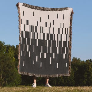 Könnte beinhalten: Eine schwarz-weiß geometrisch gemusterte Decke mit Fransen. Das Design zeigt vertikale und horizontale rechteckige Formen. Die Decke wird vor blauem Himmel und grünen Bäumen gehalten.