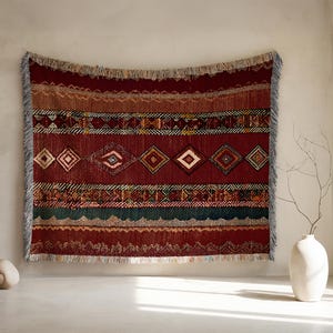 May include: A woven wall hanging with a geometric pattern in shades of red, brown, and green. The rectangular tapestry features diamond and rectangular shapes, with a fringed edge. The wall hanging is displayed against a neutral wall.