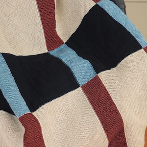 May include: A close-up of a woven blanket with a patchwork design. The blanket features squares of beige, black, burgundy, and light blue. The texture of the fabric is visible, suggesting a cozy and handmade item.