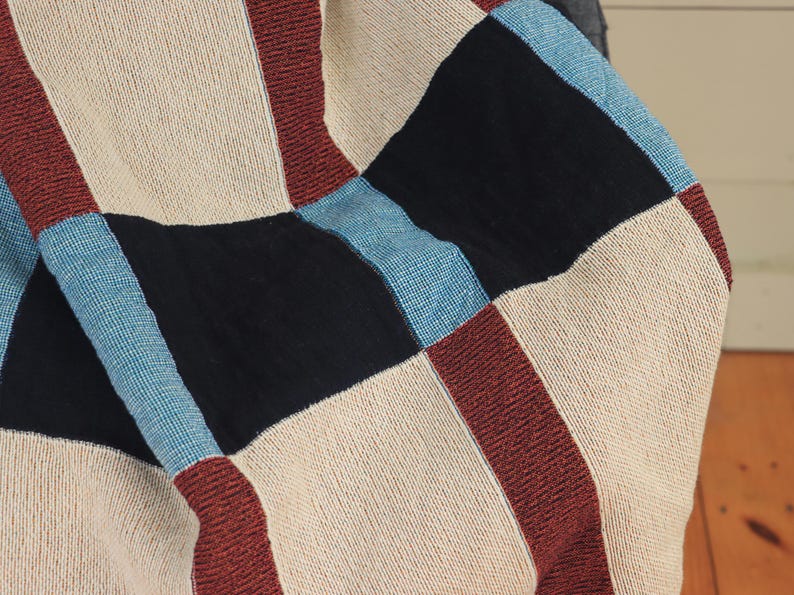 May include: A close-up of a woven blanket with a patchwork design. The blanket features squares of beige, black, burgundy, and light blue. The texture of the fabric is visible, suggesting a cozy and handmade item.