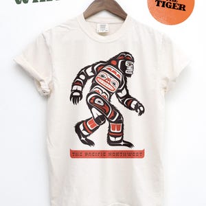 Puede incluir: Una camiseta blanca con un gr&aacute;fico de un Sasquatch estilizado en rojo, negro y azul. El gr&aacute;fico est&aacute; en el estilo del arte ind&iacute;gena de la costa noroeste. El texto "The Pacific Northwest" est&aacute; impreso debajo del gr&aacute;fico.