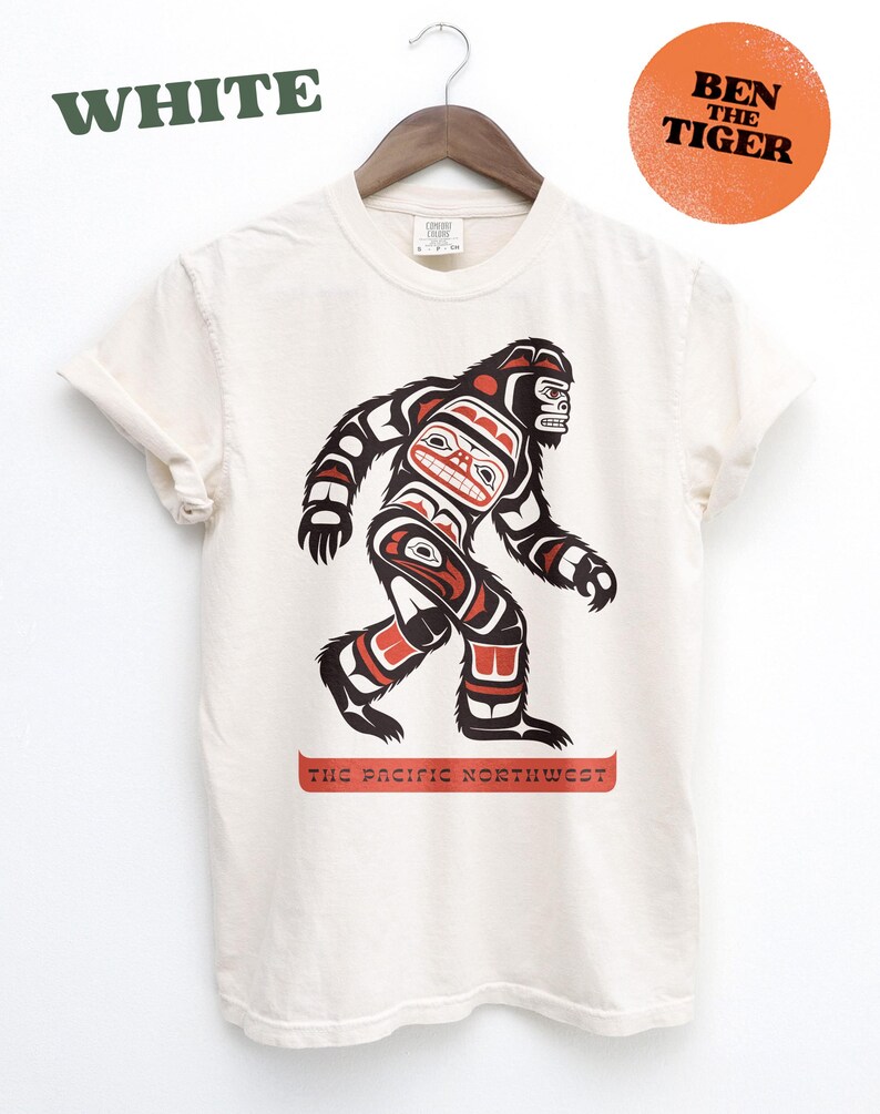 Puede incluir: Una camiseta blanca con un gr&aacute;fico de un Sasquatch estilizado en rojo, negro y azul. El gr&aacute;fico est&aacute; en el estilo del arte ind&iacute;gena de la costa noroeste. El texto "The Pacific Northwest" est&aacute; impreso debajo del gr&aacute;fico.