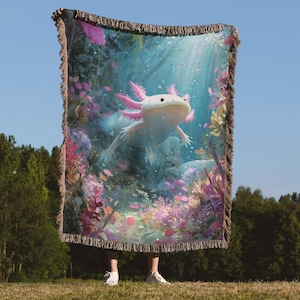 May include: A woven throw blanket depicting an axolotl in an underwater scene with pink and purple flowers. The blanket has a fringed edge. The blanket is held up against a blue sky background.