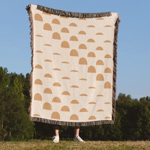 May include: A beige woven throw blanket with a repeating pattern of brown half-circle shapes. The blanket has a fringed edge in shades of brown and black. The blanket is held up against a blue sky background.