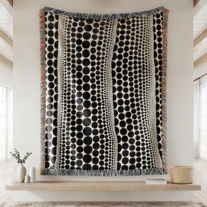 May include: Black and white woven throw blanket with a repeating dot pattern. The design features varying sizes of black circles on a cream background, with fringed edges. The blanket is displayed on a wall.