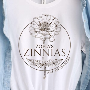 May include: White t-shirt featuring a detailed brown illustration of a zinnia flower within a circle. The text "Zofia's Zinnias" is centered below the flower, with "ALS Awareness" written along the circle's edge. The shirt is worn with a denim jacket.