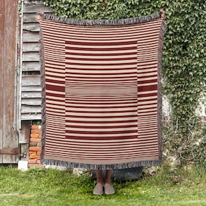 May include: A burgundy and cream striped throw blanket with a fringed edge. The blanket is held up, revealing a central rectangular design with horizontal stripes. The background includes greenery and a wooden structure. The blanket's dimensions are not visible.