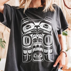 Puede incluir: Camiseta gris oscuro con un gráfico blanco de un diseño de tótem estilizado. El diseño incluye una figura central grande con elementos simétricos y una composición detallada en capas. La camiseta tiene mangas cortas y un corte holgado.
