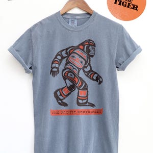 Puede incluir: Una camiseta azul jean con un gr&aacute;fico de un Sasquatch estilizado al estilo del arte ind&iacute;gena de la costa noroeste. El texto "The Pacific Northwest" est&aacute; impreso debajo del gr&aacute;fico. El texto "Ben The Tiger" est&aacute; impreso en un c&iacute;rculo de color melocot&oacute;n en la esquina superior derecha.