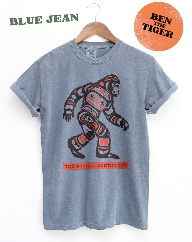 Puede incluir: Una camiseta azul jean con un gr&aacute;fico de un Sasquatch estilizado al estilo del arte ind&iacute;gena de la costa noroeste. El texto "The Pacific Northwest" est&aacute; impreso debajo del gr&aacute;fico. El texto "Ben The Tiger" est&aacute; impreso en un c&iacute;rculo de color melocot&oacute;n en la esquina superior derecha.