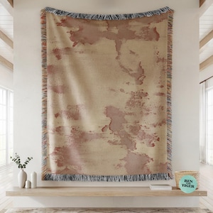 May include: A large, rectangular throw blanket with a beige background and abstract, reddish-brown blotches. The blanket has a fringed edge with a mix of colors. The blanket is hanging on a white wall.