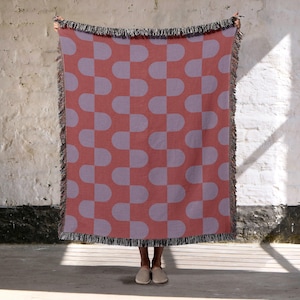 May include: A red and lavender throw blanket with a repeating pattern of rounded rectangles. The blanket has a fringed edge and is held up against a white brick wall.
