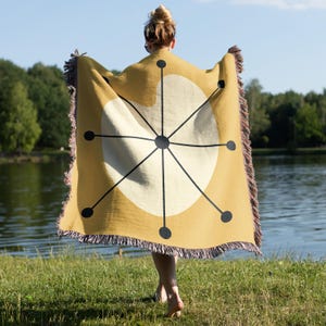 May include: Mustard yellow throw blanket with a white circle and black geometric design. The blanket has a fringed edge with shades of pink and brown. The person holding the blanket is standing near a lake.