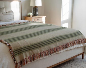 Puede incluir: Una manta a rayas beige y verde con un borde con flecos está sobre una cama. La manta presenta rayas verde oscuro y una franja con acentos rojos, naranjas y beige. La cama tiene un cabecero de color claro y un marco de madera.