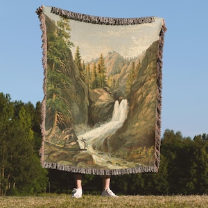 May include: A woven throw blanket featuring a landscape scene with a waterfall, trees, and mountains. The blanket has a fringed edge and is held up against a blue sky. The color palette includes greens, browns, and beige.