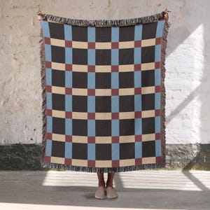 Peut inclure: Un plaid tissé présentant un motif à grille répétitif composé de blocs rectangulaires gris anthracite, bleu clair et beige. La couverture possède des bords à franges en haut et en bas, avec une texture visible sur tout le tissu tissé.