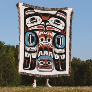 May include: A white woven blanket with a Native American-inspired design. The artwork features a central totem-like figure in black, red, and blue, with a fringed edge. The blanket is held up against a backdrop of a blue sky and green trees.