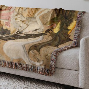 May include: Woven throw blanket featuring a medieval illustration of a knight on horseback battling a dragon. The artwork is in shades of beige, brown, and green, with a fringed edge. The blanket is draped over a light gray sofa.