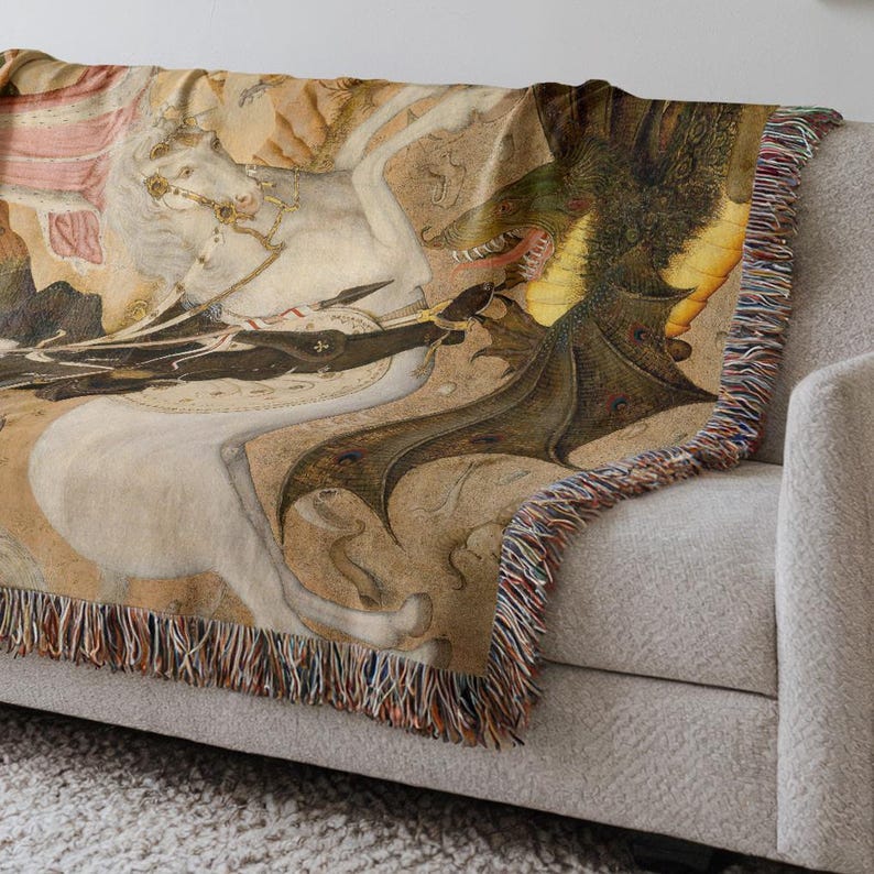 May include: Woven throw blanket featuring a medieval illustration of a knight on horseback battling a dragon. The artwork is in shades of beige, brown, and green, with a fringed edge. The blanket is draped over a light gray sofa.