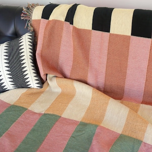 May include: A colorful throw blanket with vertical stripes in shades of peach, pink, cream, and green, draped over a sofa. A black and white patterned pillow rests on the sofa. The blanket has a fringed edge.