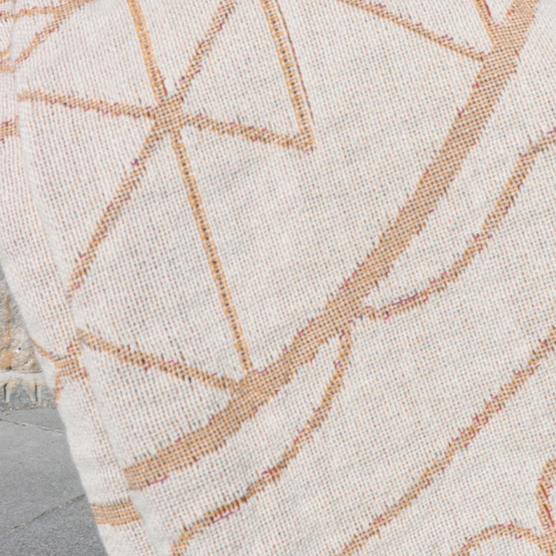 May include: Close-up of a cream-colored textile with a geometric pattern in shades of tan and brown. The design features intersecting lines and shapes, creating a modern, abstract look. The fabric has a woven texture.