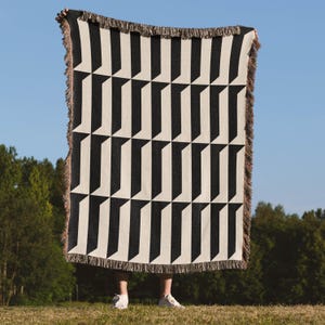 May include: A black and white geometric patterned throw blanket with a fringe border. The blanket is held up against a blue sky and green trees. The pattern consists of repeating black and white shapes.