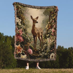 May include: A tapestry throw blanket featuring a deer in a forest scene with flowers. The blanket has a fringed edge and is held up against a blue sky. The deer has brown spots and is standing on a path.