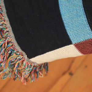 May include: Close-up of a woven throw blanket with a colorful fringe. The blanket features a black background with blue, white, and red stripes. The fringe is a mix of rainbow colors, adding a vibrant touch to the design.