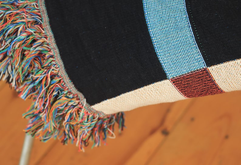 May include: Close-up of a woven throw blanket with a colorful fringe. The blanket features a black background with blue, white, and red stripes. The fringe is a mix of rainbow colors, adding a vibrant touch to the design.