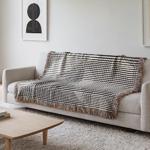 May include: A black and white geometric patterned throw blanket draped over a light gray sofa. The blanket features a repeating pattern of curved lines and stripes. The edges of the blanket have a fringe.