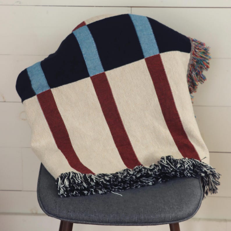May include: A woven blanket with a cream background and vertical burgundy stripes. The blanket has a navy blue and light blue square pattern at the top and a fringe trim. The blanket is draped over a gray chair.