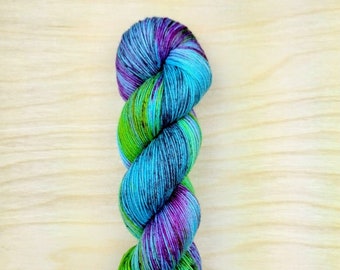May include: A skein of multi-coloured yarn, featuring blue, purple, and green hues. The yarn is twisted, displaying a gradient effect from green to purple. The yarn is presented on a light wooden surface, suitable for knitting or crochet projects.