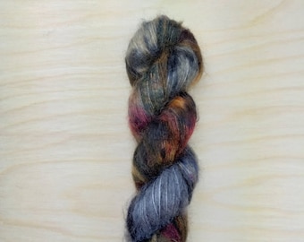 May include: A twisted skein of yarn in shades of gray, brown, and burgundy. The yarn has a soft, fluffy texture and appears to be a blend of fibers. The colors transition throughout the skein, creating a visually interesting effect. The yarn is displayed against a light wood background.