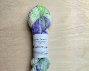 May include: A skein of Lavender Fog hand-dyed yarn. The yarn is a mix of white, light green, and purple. The label reads "Blackbird Sycamore" and includes details about the yarn's composition and weight, including 100 grams.
