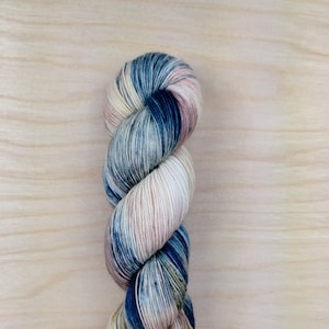 Könnte beinhalten: Eine Garnrolle in den Farben Blau, Creme und Rosa. Das Garn ist handgefärbt und hat eine gesprenkelte Optik.