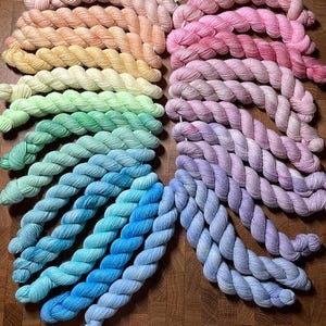 May include: A collection of twisted yarn skeins arranged in a circular gradient pattern. The colors transition through the spectrum, featuring shades of peach, yellow, light green, turquoise, blue, purple, and pink, all neatly coiled and displayed.