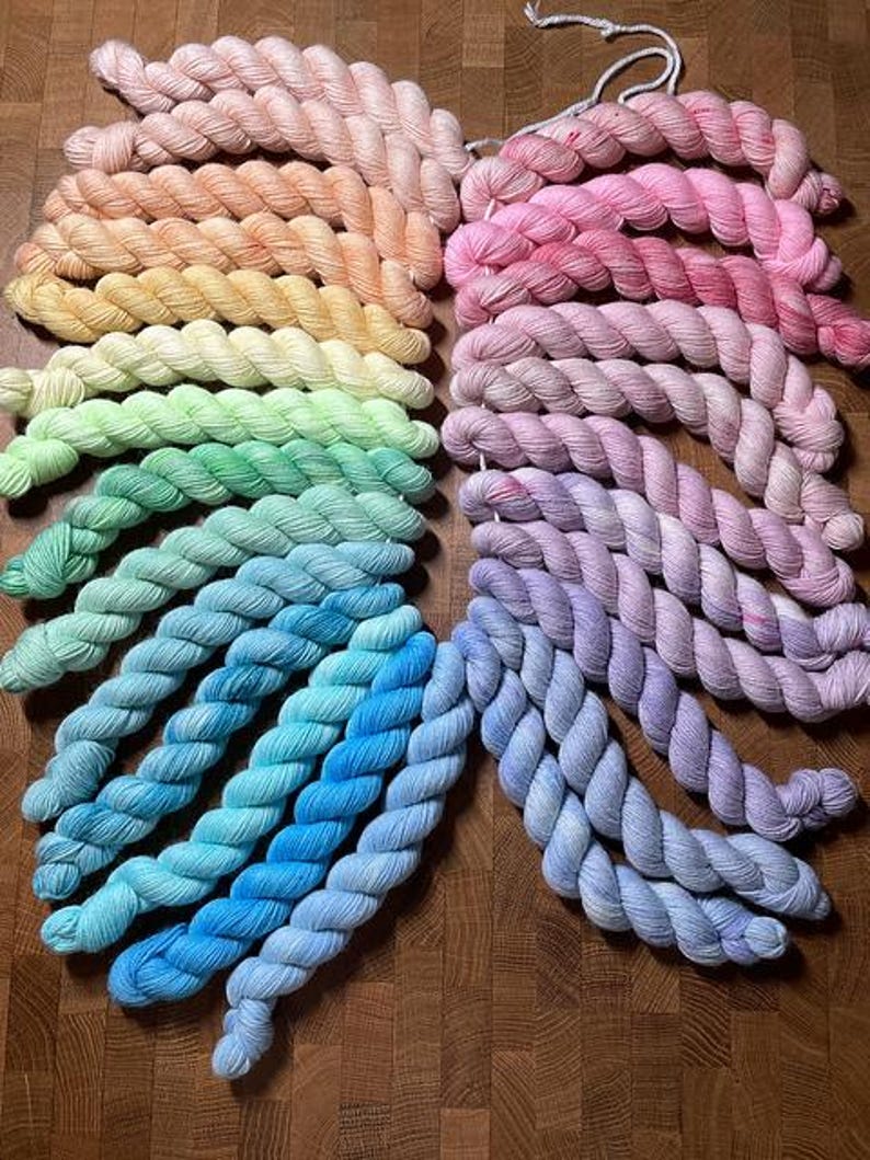 May include: A collection of twisted yarn skeins arranged in a circular gradient pattern. The colors transition through the spectrum, featuring shades of peach, yellow, light green, turquoise, blue, purple, and pink, all neatly coiled and displayed.
