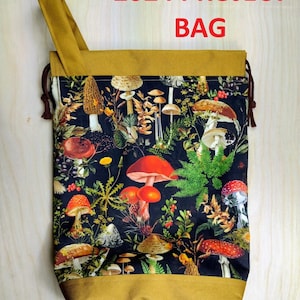 May include: A project bag with a mustard yellow top and bottom band, featuring a black fabric center printed with a colorful botanical pattern of various mushrooms, ferns, and berries. The text 2024 PROJECT BAG is printed in red above the bag.