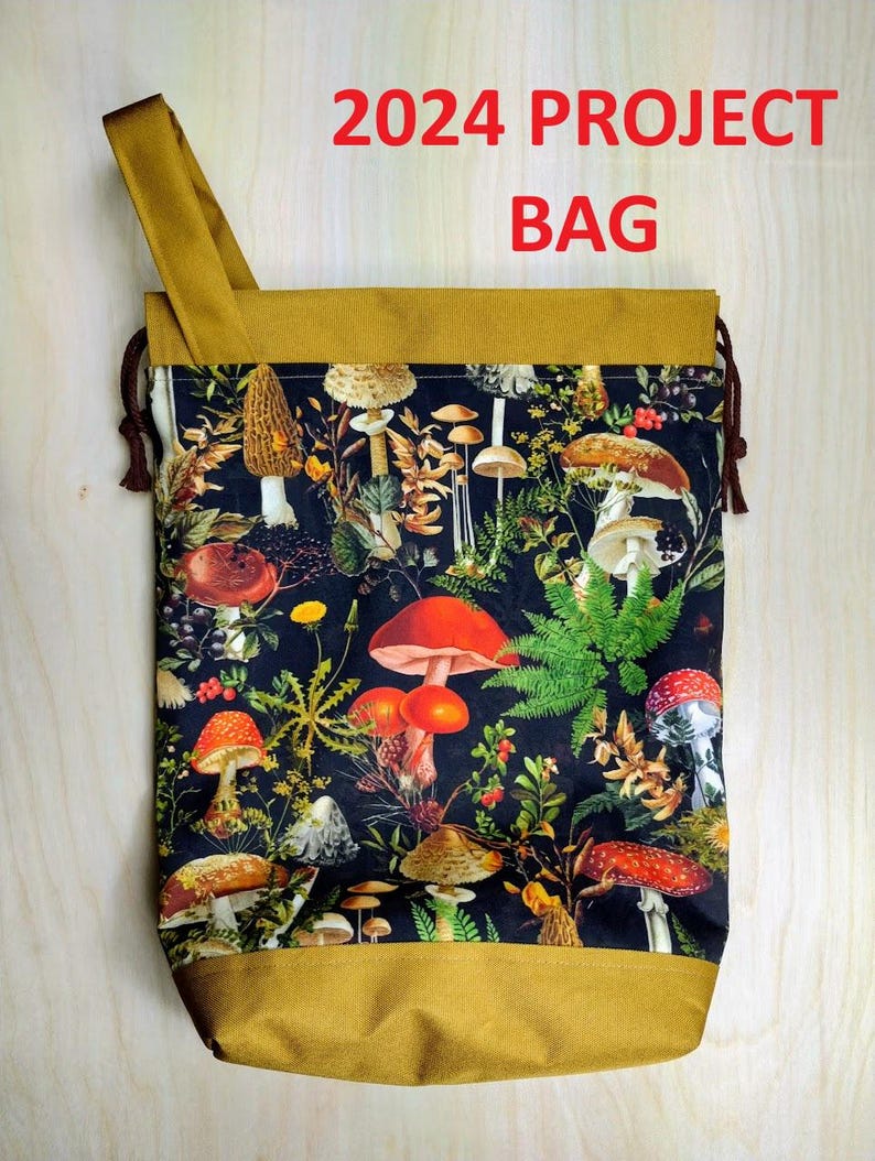 May include: A project bag with a mustard yellow top and bottom band, featuring a black fabric center printed with a colorful botanical pattern of various mushrooms, ferns, and berries. The text 2024 PROJECT BAG is printed in red above the bag.