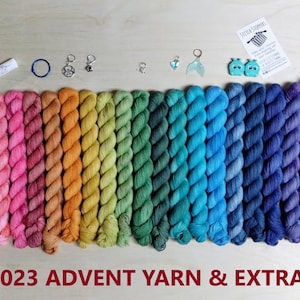 May include: A collection of twenty-four mini yarn skeins arranged in a rainbow gradient, accompanied by small metal stitch markers, a lip balm, two teal whale-shaped stitch stoppers, and a small card labeled 2023 ADVENT YARN & EXTRAS.